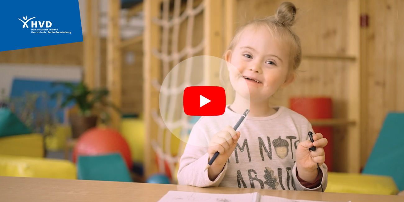 Mädchen sitzt an Spieltisch und lächelt in die Kamera, Foto verlinkt auf Video auf YouTube